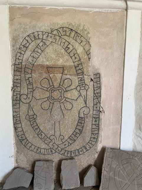 Photo of a runestone mounted in a wall. The stone is reddish, with black inscriptions inside two rune snakes, and a cross in the middle. There are a few tiny runestone fragments laying on the floor next to it. 