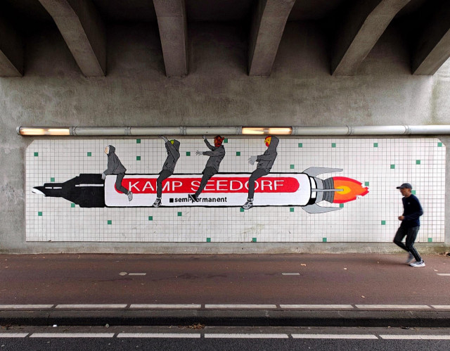 A photo showing street art under a bridge: a large paper shows figures in hoodies riding a rocket-shaped pen labeled “KAMP SEEDORF” and “semi permanent”. A person is jogging past on the road below.