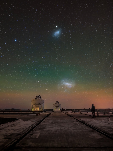 Two of ESO’s small Auxiliary Telescopes, accompanied by the Magellanic Clouds up in the night sky. The ATs are small white domes, their tops open revealing telescopes pointing at the sky. The Magellanic Clouds are look like bright, blueish clouds with stars within them. A human silhouette is visible on the right side of the picture.