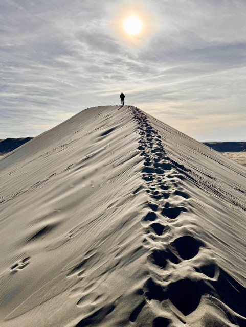 Photo of hiker standing at peak of ridge as viewed from the ridge. View is from the ridge looking toward the sun which is also in the pic. Footprints are seen in foreground on ridge.