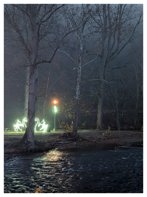 	
A misty, wooded scene at night features a glowing street lamp near leafless trees. Neon swan-shaped lights are arranged on the left side. In the foreground, water reflects the lights, creating a dreamy atmosphere under the foggy sky.