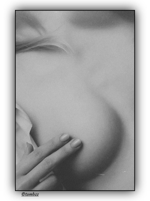 Black and white analog photo showing an intimate, sensual close-up of a woman's body. The focus is on her breast, gently covered by her fingers, with the nipple hidden in a soft, protective gesture. Light falls softly across her skin, bringing out the natural curves and quiet tension in the frame. A hint of blonde curls appears at the top, adding warmth and texture. The composition is minimal but charged, balancing between presence and concealment. The image feels calm, powerful and deeply sensitive.