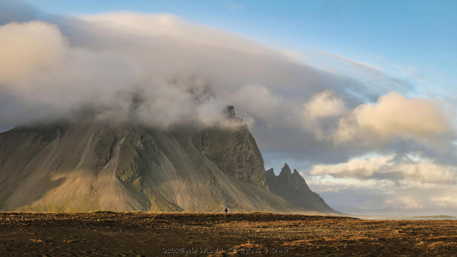 A colour photo of an impressive mountain range. The peaks are hidden behind a cap of dense cloud but blue sky can be seen above. The lower slopes of the scree are sunlit and the crags are distinct. The foreground is black sand dunes and in the centre near the bottom is a solitary figure.