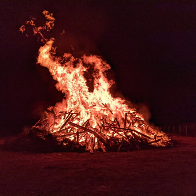 This image depicts a large bonfire burning intensely at night. The flames are vivid and dynamic, rising high above a substantial pile of wooden logs and debris. The fire illuminates the surrounding area with a warm, orange glow, contrasting sharply against the dark night sky. The scene captures the raw energy and heat of the fire, with embers and sparks visibly floating upwards. The bonfire appears to be situated in an open outdoor space.