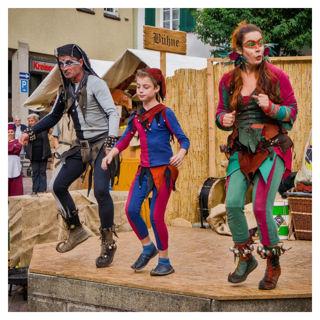 Three performers in vibrant medieval-fantasy costumes are captured mid-jump on a small wooden outdoor stage during a sunny street festival in a German town. The leftmost is an adult man dressed as a pirate or rogue, wearing a black tattered hood, white skull-like face paint around his eyes and mouth, a gray sleeveless jerkin over a light shirt, black pants with a white stripe down one leg, and rugged brown boots adorned with bells and straps, leaping forward with an intense expression. In the center, a young boy in a jester outfit sports a red pointed cap with bells, a blue-and-red split tunic and matching leggings with a black collar, and simple blue shoes, jumping with relaxed arms and a neutral look. On the right, an adult woman as an elf or fairy has long reddish-brown hair in a high ponytail with pink flowers and green antennae-like headpieces, green face paint with red dots and leaf patterns, a green-and-maroon laced corset, turquoise-and-magenta leggings with leaf details, and brown lace-up boots with bells, mid-leap with her right fist raised and a playful puckered-lip expression. The background features a light-colored building with green shutters, a wooden sign reading “Bühne” for stage, a beige canvas tent, woven reed fencing, a large drum, wicker basket, wooden crates, a few onlookers in mixed period and modern clothing, creating a festive and energetic atmosphere.
