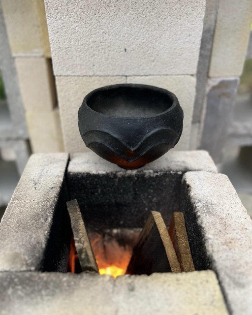 A small black bowl sits above the furnace of the rocket kiln as a fire burns below. The bowl was smoke fired to blacken it. The furnace is square and formed with off-white fire brick. Scrap would is partially visible in the burning chamber. The bowl is simple but well formed and features two scalloped decorative ridges on the exterior surface. 