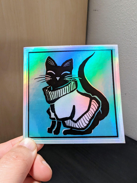 Photo of a left hand holding a square sticker. The sticker is a holographic black cat in a sweater drawing.