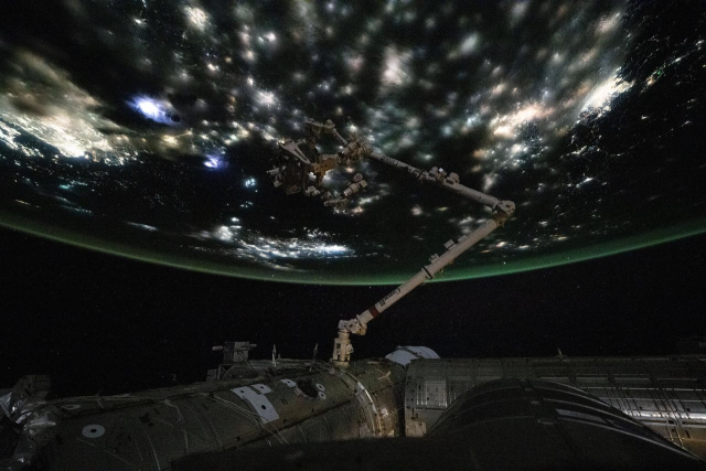 A view from the International Space Station showing part of its structure and robotic arm silhouetted against Earth at night. The planet’s surface glows with lightning flashes and illuminated cities beneath a faint green atmospheric layer along the horizon.