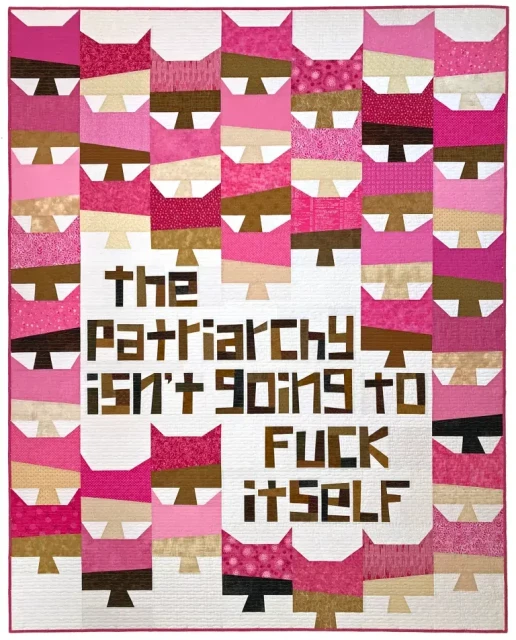 Full view of a pussy hat-themed quilt with columns of stylized blocks resembling a neck and face wearing a pussy hat. The blocks are wonkily pieced from scrappy pinks (the hats) with peach, beige, brown and black fabrics for the body parts. An asymmetric section of the quilt near the bottom center features improv pieced letters instead of pussy hat blocks; it reads "the patriarchy isn't going to fuck itself" in scrappy brown fabrics on a white background.