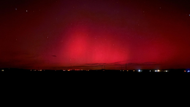Aurora in the night sky glows red above lights dotting a black horizon. 