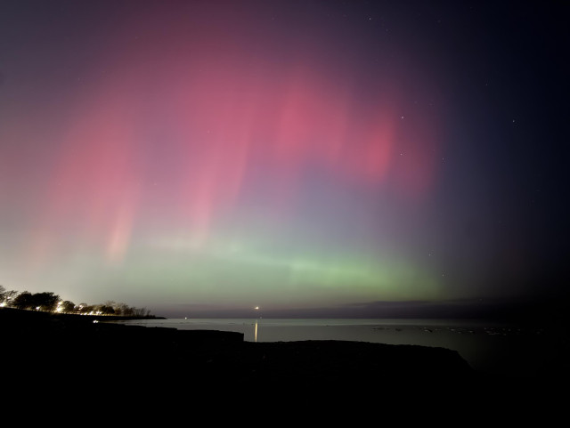 Bright greens and reds light up the sky over Lake Michigan. A small light shines on the horizon. Some trees are visible on the lower left and lead into darkness on the bottom of the frame. 