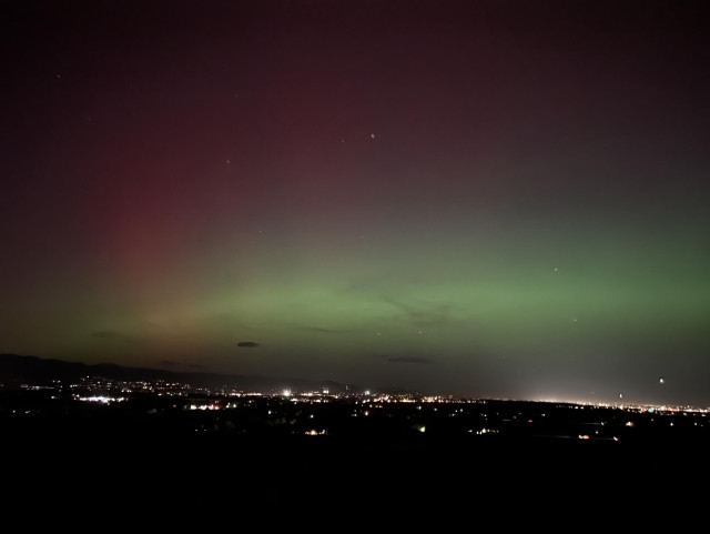 The image showcases a vibrant night sky illuminated by an aurora, displaying shades of green and purple. Below, a cityscape with lights can be seen against the dark landscape.
