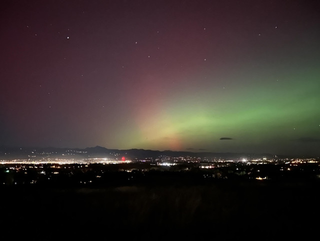 A night sky displaying auroras in vibrant shades of green and purple over a cityscape, with lights from buildings and mountains visible in the foreground.