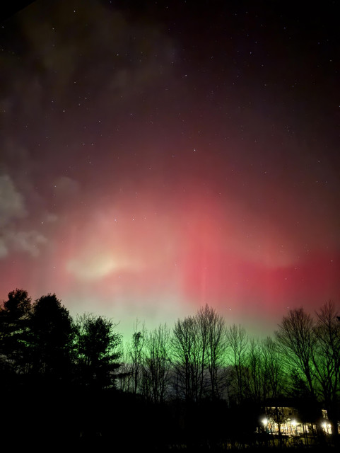 photo of aurora as clouds start moving in, very green along the horizon and through trees, while above it is shades of red fading into the dark night sky full of stars. In lower right a large house with lights wrapped around its perimeter glows warmly. 