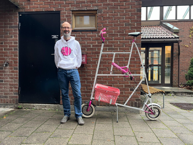 Middle aged, beared, white man looking at the camera and smiling. He is wearing a white hoodie with a pink, piggie bank logo (Bónus) and blue jeans. He is standing next to a pink and white tallbike with tiny wheels. It also has a pink basket.