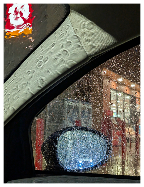 Rain-speckled car window seen from inside at night; blurred gas station lights and signs appear outside. A glowing blue car headlight is reflected in the side mirror, and raindrops cover both the glass and door frame. A red and white sign is partially visible above.