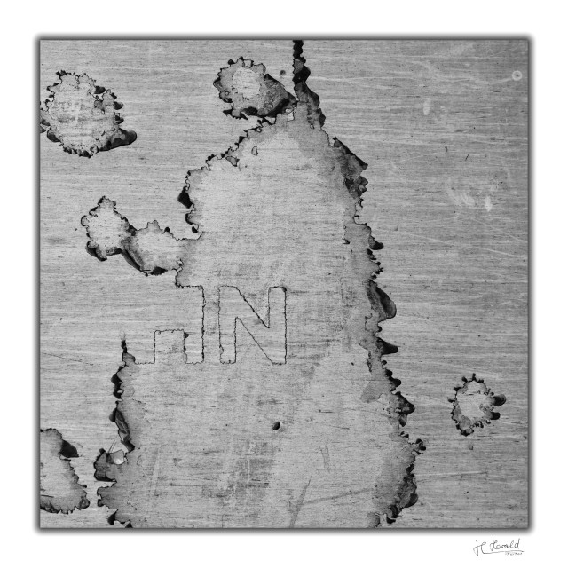 Square black-and-white image of a peeling sticker on a train. The resulting patterns look like a map of islands. In the centre, half an ‘H’ and an ‘N’ can be seen. The dirt stuck to it has been smudged horizontally by the airstream. 