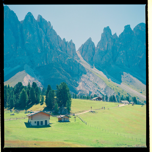 A sun drenched alpine meadow features rustic wooden cabins and a winding path, set against towering, jagged mountain peaks. Lush green grass, scattered trees, and distant hikers create a serene, pastoral scene under a clear blue sky.