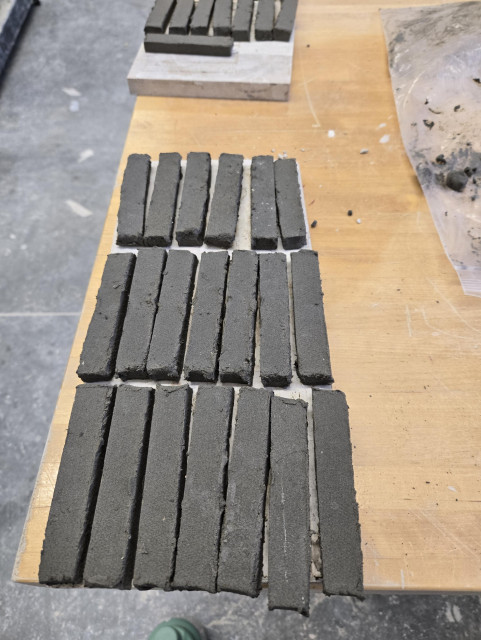 About two dozen dark gray bars of clay lined up in rows, sitting on a wooden table in what seems to be an institutional setting (school lab).