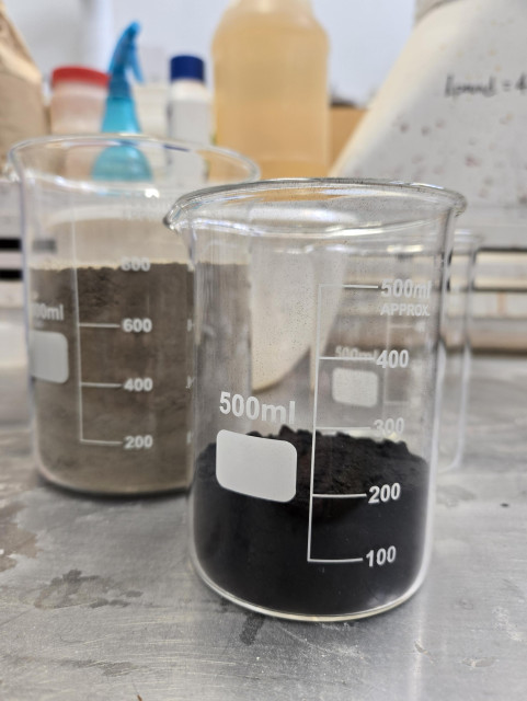 Two graduated cylinders. One filled with dry, pulverized gray clay and the other filled with black pulverized burned sea sponge (sponge spicules).