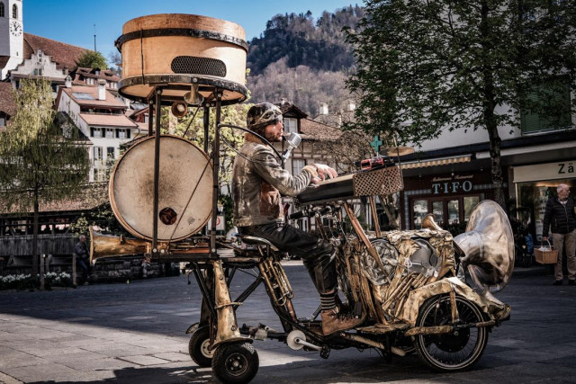 EIn Musiker auf einer Art Steampunk-One-Man-Band-Musiziergefährt. Das Teil wird mit Pedalen angetrieben, hat diverse Lautsprecher, eine Pauke, Trompetenhörner, Mikrofon, Piano und ganz vorne eine Tuba. Auch irgendwo im Bild aber nicht in Aktion: eine Rauch- und eine Seifenblasenmaschine .