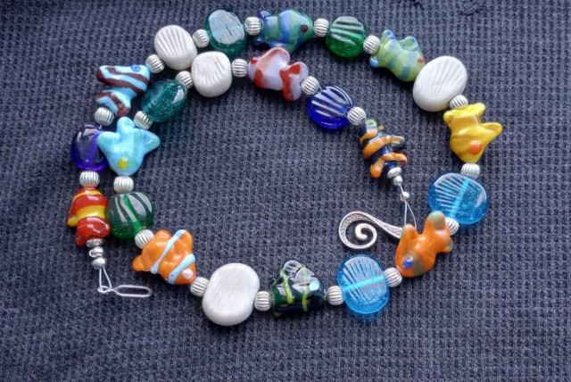 Photo of a handmade lampwork glass bead necklace in a wide range of colors, featuring beads sculpted to look like tropical fish and seashells, set in a spiral pattern against a dark blue fabric background.