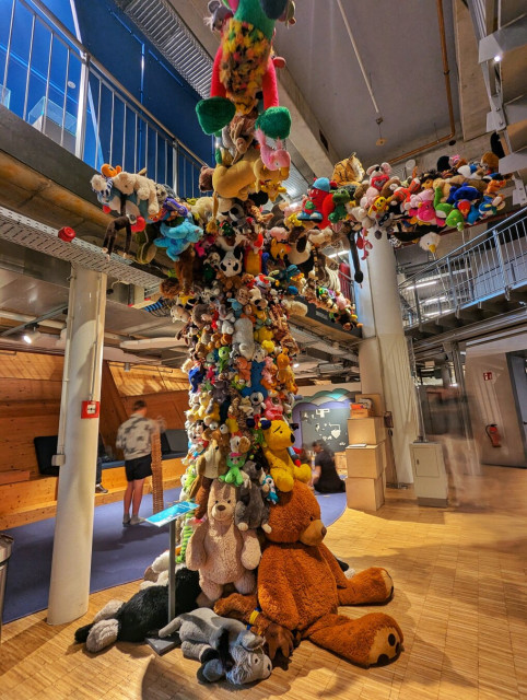 Interior view of a spacious, modern building featuring a towering, whimsical art installation made entirely of plush toys. The installation resembles a tree or column, densely packed with a vibrant assortment of stuffed animals, including teddy bears, dogs, cats, and various other creatures. The toys are stacked and arranged to create a visually striking and colourful display that stretches from the floor to the ceiling. The surrounding area appears to be an open, multi-level space with wooden flooring, metal railings, and industrial-style lighting. In the background, a few people are visible, adding a sense of scale to the impressive installation.