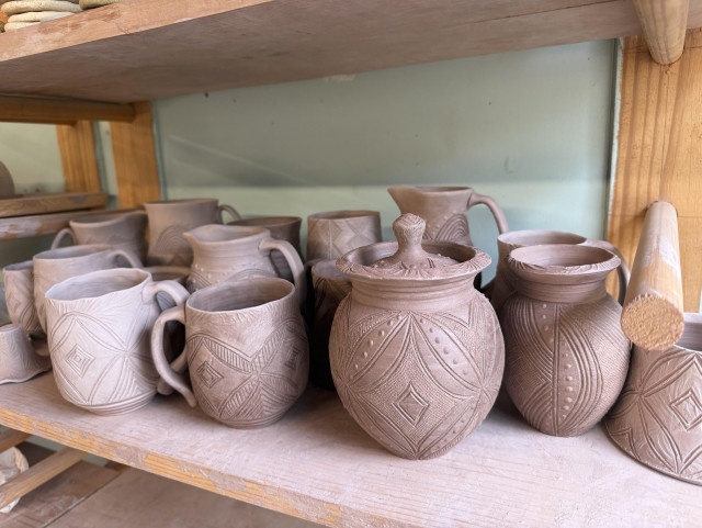 A small batch of bone-dry pots sitting on a wooden shelf. Intricately decorated jugs, jars, vases and vases are clustered together with a light blue wall in the background. 