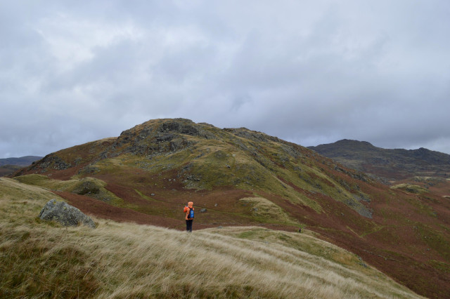 a small figure in fluorescent orange jacket stands on a windswept grassy hill, with a craggy semi-mountainous horizon behind. the sky is wild and there's no one else in sight.