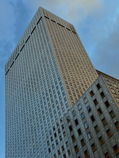 A color photograph in portrait orientation that captures the dramatic upward view of a large, modern skyscraper in midtown Manhattan against a partly cloudy blue sky. The building occupies most of the frame, towering diagonally from the lower right toward the upper left. The main structure is characterized by its sheer verticality and a highly textured, grid-like facade composed of numerous small, repeated rectangular windows and metal panels. The material is a silvery-gray muted metallic color, reflecting the ambient light unevenly.