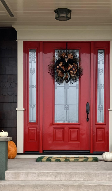 A red front door with art deco style glass panels. A seasonal wreath hangs from a wreath hanger. Some pumpkins are still on the porch to the side of the door. 