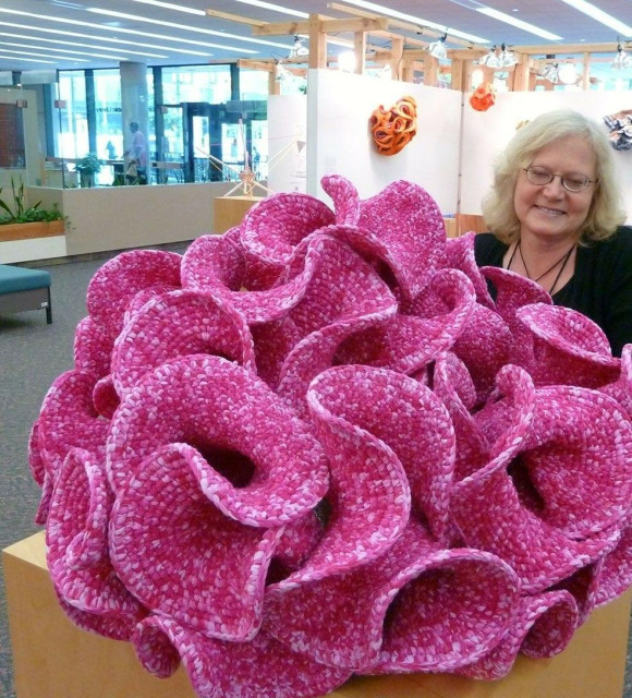 Photo of a white woman standing by a table on which a large pink crocheted organic looking piece rests