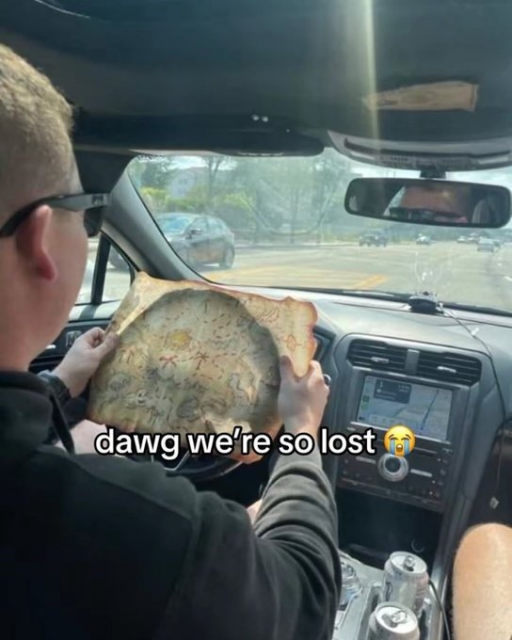 A humorous photo taken from inside a car, looking over the driver's shoulder. The driver is holding up a large, weathered parchment map that resembles a fictional treasure map rather than a navigational tool. Through the windshield, a standard paved road with traffic and trees is visible. White text overlaid on the image reads, "dawg we're so lost 😭".