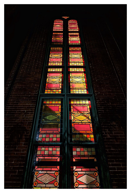 Nighttime photo of a multistory stained glass window in a brick facade, taken from street level looking up. The window is arched at the top and consists of two adjacent columns of tall rectangular panels, each with an abstract design of geometric shapes in hues of red and orange. The window is lit from within, glowing in the nighttime darkness, and the diagonal shadow of a staircase and banister can be seen against the lower panels.