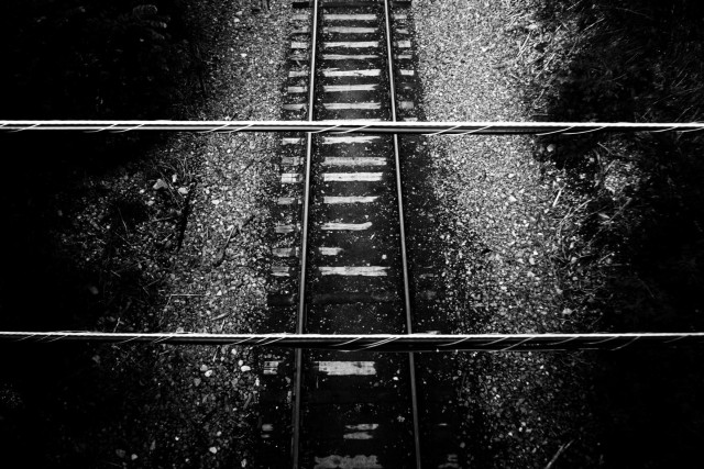 The photographer stands on a bridge looking down onto train tracks heading vertically a pair of wires are seen crossing horizontally. This is a very high contrast black and white image.