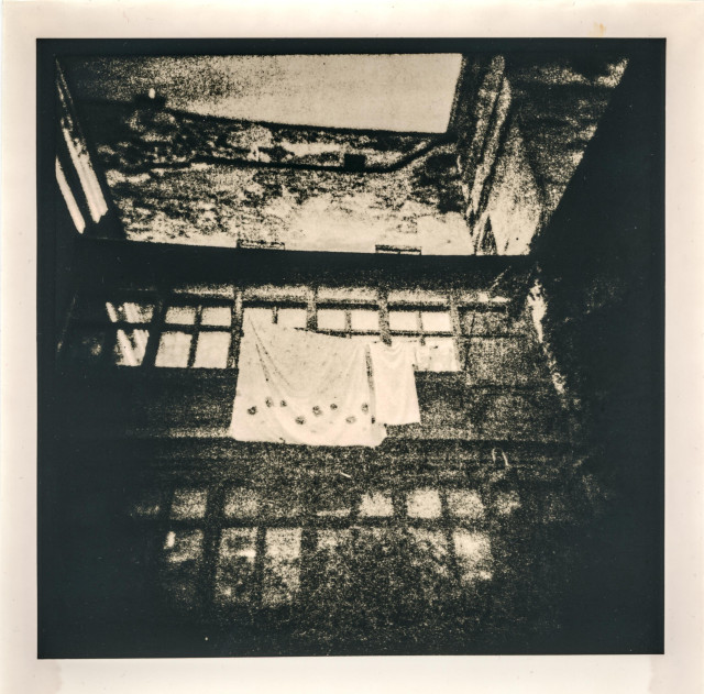 This lith print captures the atmosphere of an old Lviv courtyard, rendered in gritty, high-contrast tones that emphasize texture and decay. The composition looks almost like a reflection or a memory—distorted, soft at the edges, and saturated with grain. Laundry hangs across a row of worn windows, its pale fabric glowing against the deep blacks surrounding it. Above, the rough surfaces of aging walls dissolve into abstract patterns, giving the scene a dreamlike, slightly haunted quality. The print feels both intimate and distant, as if the courtyard is being recalled from a fragment of time rather than seen directly