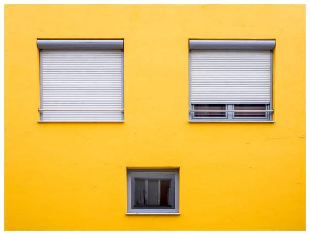 Ausschnitt einer Hauswand, dottergelb. Darauf drei Fenster, zwei oben, eins unten - jeweils über die Breite ausgemittelt. Die oberen zwei etwas größer, Rollos geschossen. Das untere ohne Rollo.