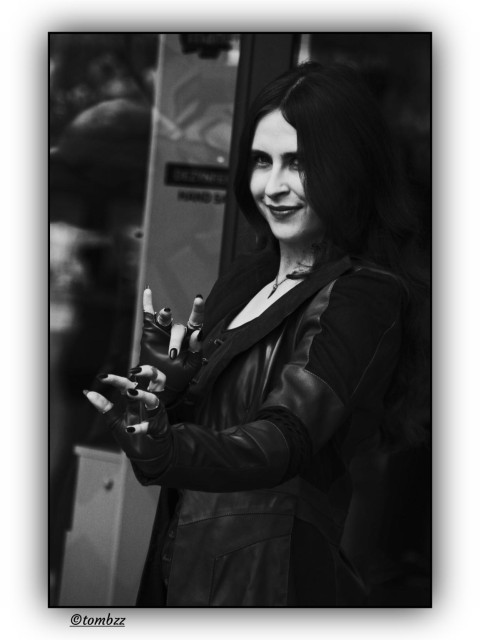 This black-and-white photo shows a young woman dressed as Scarlet Witch from the Marvel universe. Her long dark hair falls loosely over her shoulders. She’s wearing a shiny, leather-like jacket with decorative stitching and dramatic cuts. Her hands are stretched toward the camera, fingers spread wide, with long black-painted nails that add to the theatrical feel. Her makeup is bold, especially around the eyes, and her expression is confident with a hint of mischief. The background includes part of a door or shop window with reflections and blurry signage, suggesting an urban setting. The whole frame feels stylized and cinematic, with a strong, magical energy that fits the character perfectly.