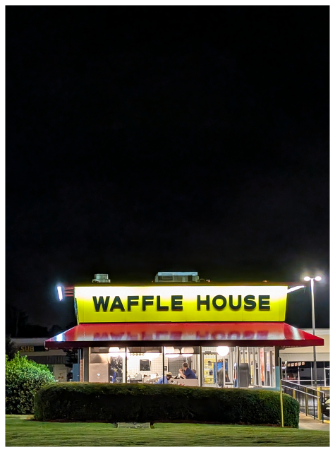 A brightly lit Waffle House restaurant stands out at night, with a yellow sign and bold black letters. Red trim decorates the awning. The windows reveal people inside, and the building is surrounded by bushes and a dark, cloudy sky.