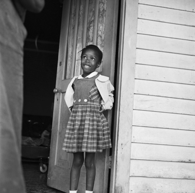 Black and white photo of Ruby Bridges as a young schoolgirl, circa 1960. She is a beautiful, bright-eyed Black child. She stands with her back against an open door, her eyes turned up, smiling.