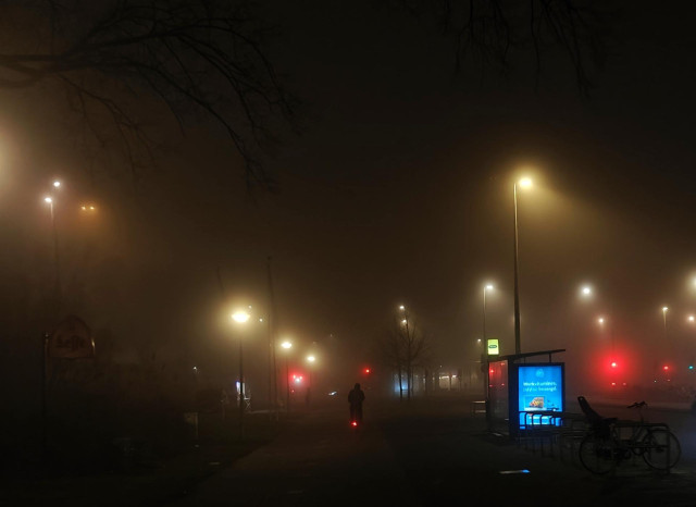 A photo of a misty street at night with streetlights and traffic lights glowing and a person cycling in the distance.