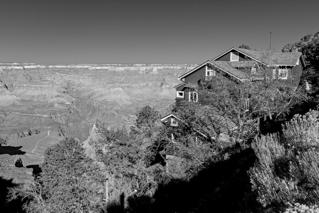A black and white landscape photo of the Grand Canyon. The view shows the Grand Canyon at center left under a dark gray sky achieved through digital processing as though a red filter had been used. The canyon is wide, deep, and very rugged. A small part of the Bright Angel Trail is seen in the lower left. On the right is a multistory building perched on the edge of the canyon. It has dark gray walls and white trim. It probably has five or maybe six levels. Trees surround the building and cut across in the foreground of the photo from the lower left to upper right.