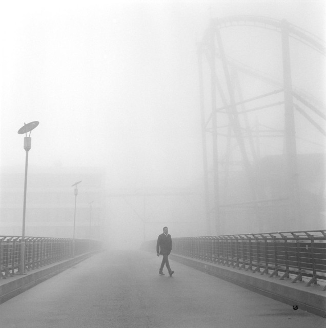 Ein Mann geht alleine über eine Brücke, es ist sehr dunstig. Im Hintergrund sind Gebäude noch zu erkennen. 