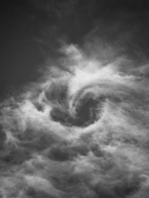 Black and white portrait format image showing a swirling vortex of wispy white clouds set against a dark sky