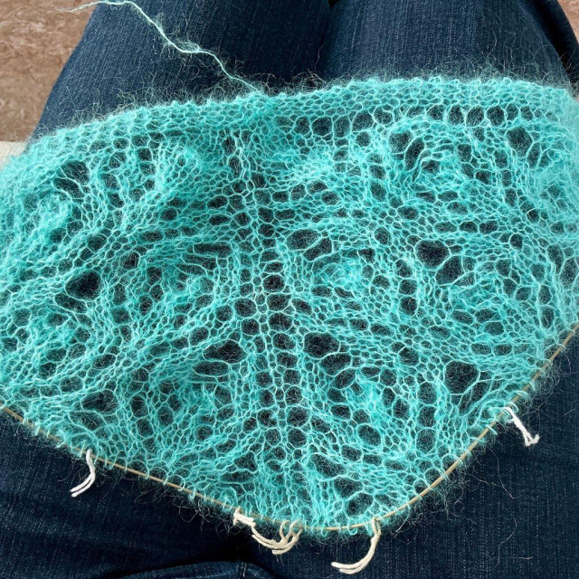Flat lay view of a bright turquoise lace shawl in kid silk yarn on the lap of someone wearing dark blue jeans.