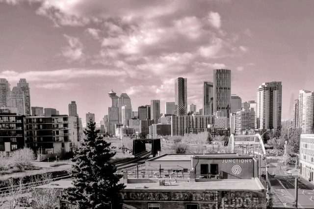 A black and white photograph depicts a Calgary cityscape on a sunny Autumn day, with a foreground of low-rise buildings and a railway line, and a background of taller skyscrapers.

Visible on the building in the foreground is signage that reads “JUNCTION 9” with a circular logo.

Photo taken from the vantage point of the Esker Contemporary Art Gallery in Calgary's Inglewood neighbourhood. 