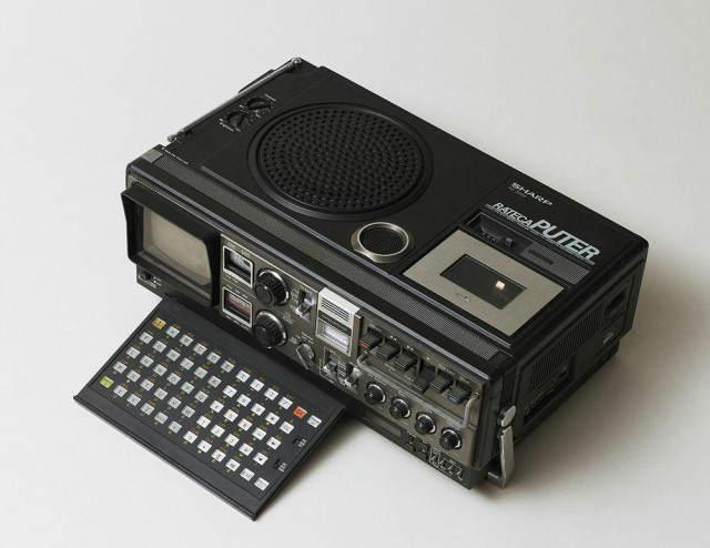 The Sharp Rateca Puter. Up top, left to right: a collapsable TV/radio antenna, brightness and contrast dials, a giant speaker, a smaller speaker, and input for the tapedeck, with the play counters that were more common on the day. 

Face, left to right: A 4.5 inch CRT (behind a plastic guard), VHF/UHF and radio dials, channel and band selectors (TV/COMP, FM, Shortwave, and Middlewave), the "RATECA PUTER' logo on the battery gauge, AFC switch, mode selector, tape deck controls, audio controls for balance, bass, treble, and volume, and a mollyguard to prevent dials and switches from being mashed (the mollyguard being a signature of Sharp's boomboxes of the time). 

Face, low: the collapsable carrying handle that doubles as a stand; computer power button; slide-out chiclet keyboard based on Sharp calculators of the day, with number pad; an LED clock, clock controls, alarm controls. 

Right side: mic jacks, earphone jacks in two different sizes, beat cut switch, fan exhaust, DC and AC power plugs, and a printer port (!).