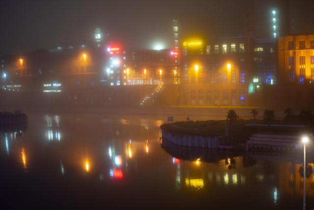 Farbfoto im Querformat. Nächtlicher Blick über das Wasser des Tempelhofer Hafens auf bunte beleuchtete Fabrikgebäude im Nebel, die Lichter spiegeln sich im Wasser. Alles ist neblig verwischt.