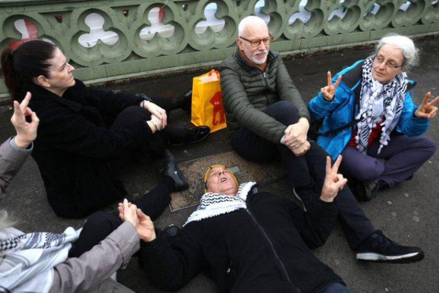 A small group of protesters sitting cross-legged on the ground.  A few are doing the peace sign, and are wearing keffiyehs.  One is lying down on the pavement.