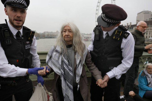 A lady with long white hair, wearing a raincoat and a keffiyeh, is being taken away by two police officers.